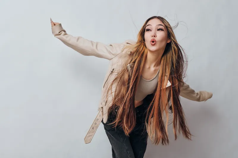 Young woman with long braided hair wearing jacket and jeans posing against white background. Female model expressing playful attitude and energetic mood in studio. Beauty, fashion and hairstyle