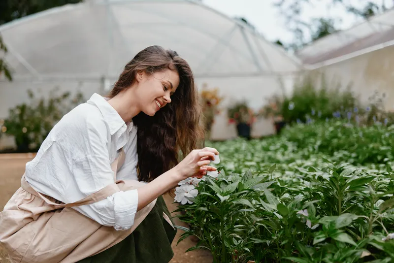Young woman tending to flowers in a greenhouse, showcasing passion for gardening, wearing casual attire and smiling, surrounded by vibrant greenery