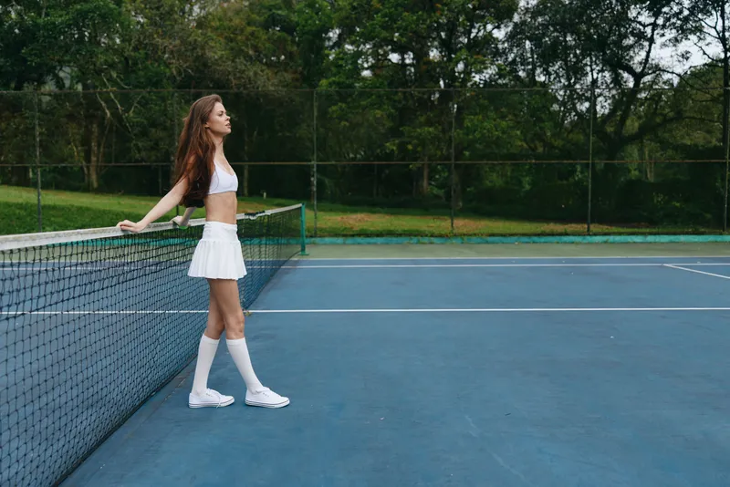 Young athletic woman in white tennis outfit stands confidently on the court, showcasing her sporty style amidst a vibrant green background, capturing the essence of sport