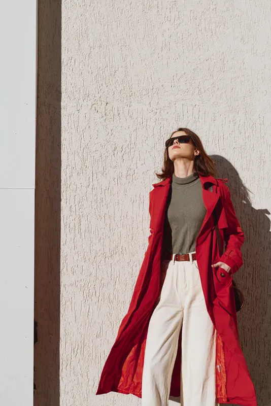 Stylish woman in a bold red trench stands against a textured wall, grey turtleneck and white pants, sunglasses on, confident pose in bright sunlight