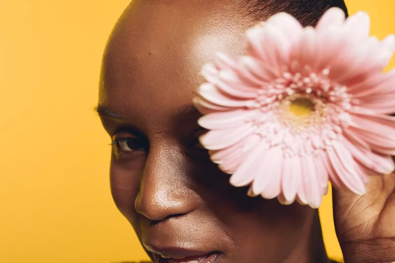 Close-up of a smiling African woman with dark skin holding a pink flower near her face on yellow background. Concept of natural beauty, floral, skincare, and wellness.