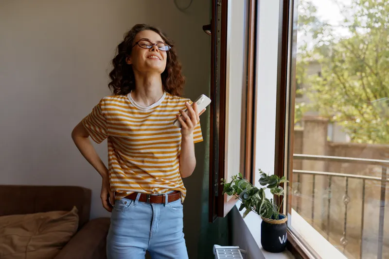 Smiling woman with curly hair wearing a striped shirt stands by a sunlit window at home, holding a phone. Casual lifestyle scene highlighting comfort, daily routine, and wellbeing. Everyday.