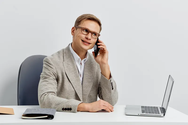 young smiling caucasian man talking phone laptop casual blazer sit at modern white table remote work digital nomad concept