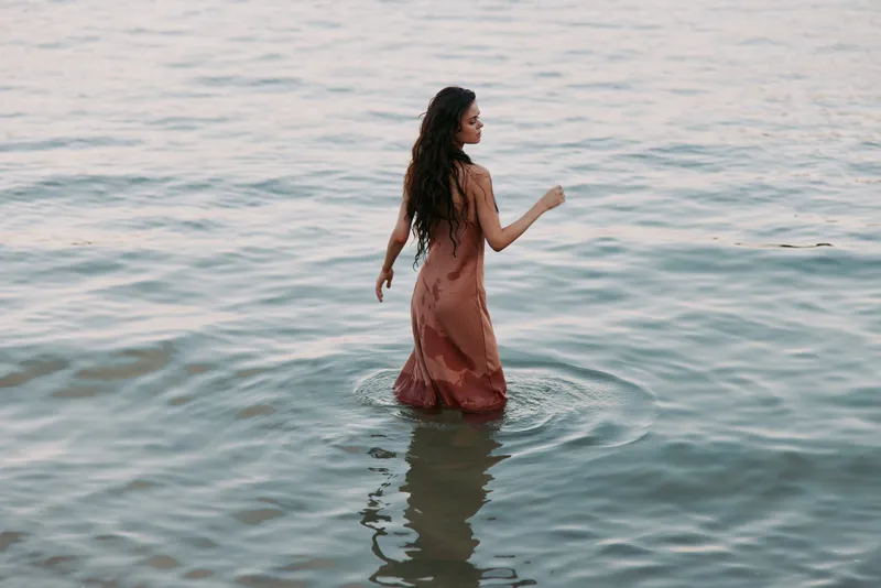 A woman in a soft pink dress walks waist-deep through calm sea water, her silhouette gliding with the tide as sunlight glints on gentle waves, creating a tranquil scene.