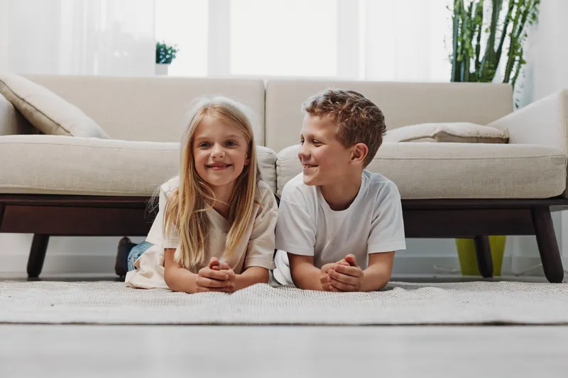 Happy children boy and girl lying on carpet in living room smiling and looking at each other. Cozy home interior background with sofa and plants. Concept of friendship and childhood.