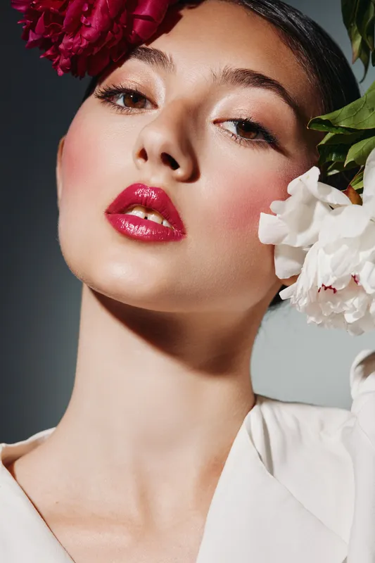 Close-up portrait of a young woman with vibrant makeup and flowers in hair, showcasing beauty, fashion, and creative styling against a neutral backdrop.