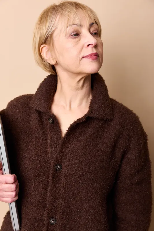 Older woman portrait holding magazine, wearing brown fleece coat, looking sideways, calm mood, studio lighting on beige background, wellness and home reading