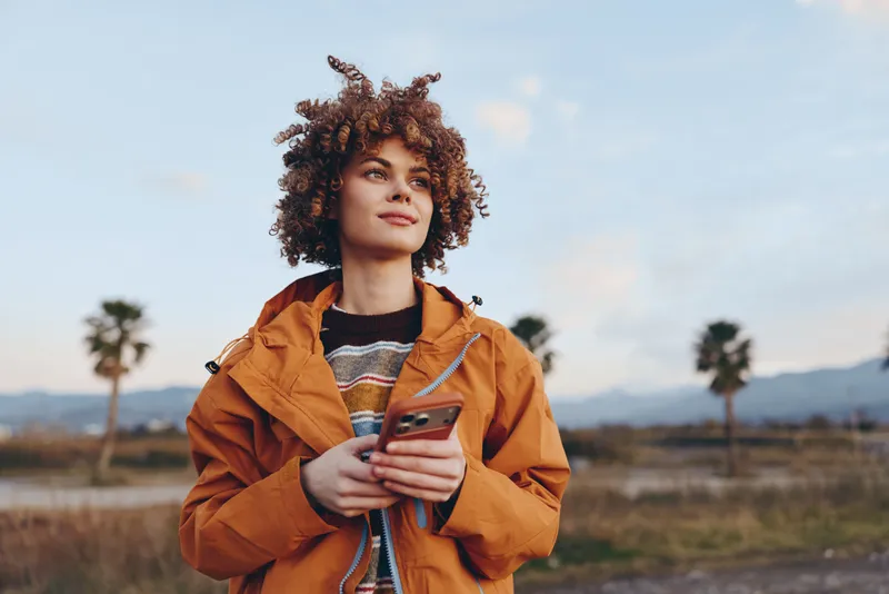 Woman lifestyle outdoors, curly hair, orange jacket, smiling, sunset, holding smartphone in hand, relaxed portrait with warm sunlit light, open field and palm trees in background