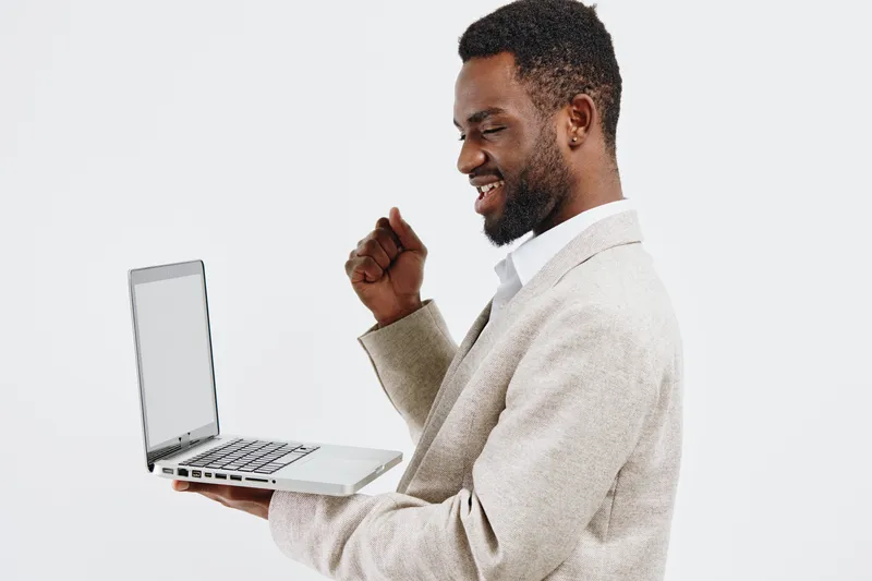 Confident smiling man holding a laptop close to chest, gesturing enthusiastically, wearing beige sweater over white shirt, standing against plain white background, studio shot, lifestyle concept, tech