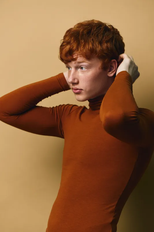 Young redhead man with freckles wearing brown turtleneck adjusting hair, looking sideways on beige background with thoughtful expression and casual style indoors.