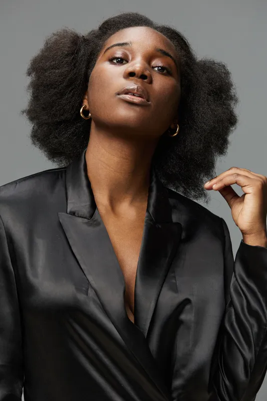 Confident female model in a sleek black blazer showcasing natural hair, exuding elegance and empowerment against a minimalist gray background