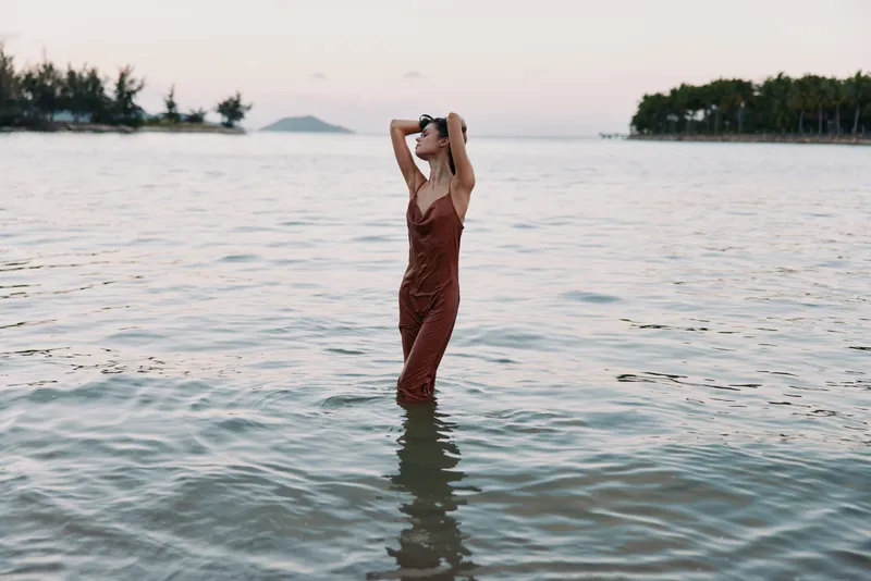 A solitary woman stands in shallow sea wearing a rust red dress, hands to hair, calm horizon, soft light, tranquil mood, coastal dusk scene.