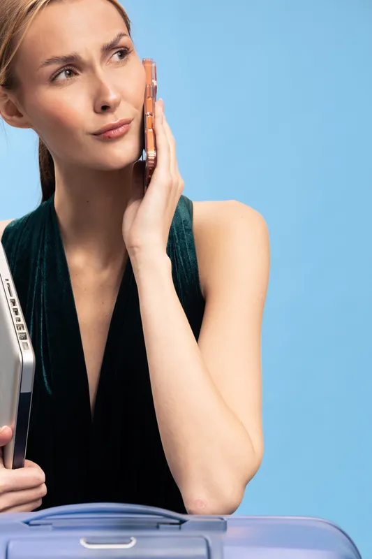 Young woman in black top holds laptop and suitcase while talking on phone with thoughtful expression. Blue background, concept of travel, communication, work and planning.