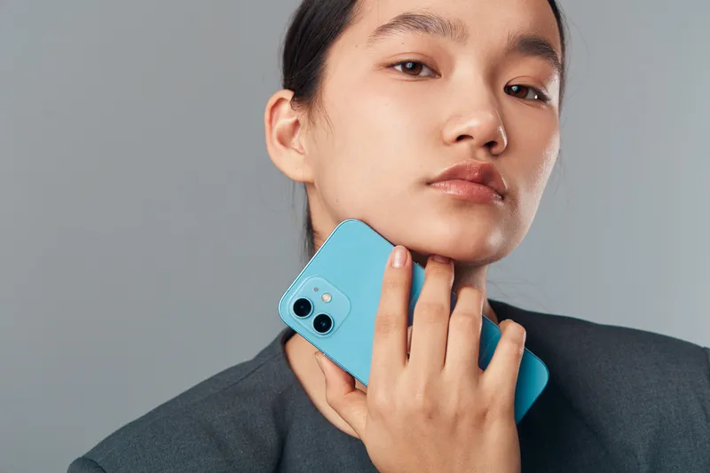Young woman holding blue smartphone near neck with serious expression, wearing dark jacket and posing against gray background. Closeup portrait with clean makeup and neat hairstyle.