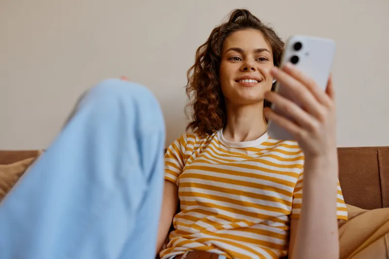 A smiling young woman sits on a sofa at home, wearing a striped tee and jeans, taking a selfie with a smartphone. A casual lifestyle moment showcasing home comfort and personal creativity.