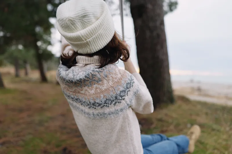 Cozy moment outdoors with a person in a knit sweater and beanie, sitting by trees in a park and gazing at a calm shoreline on a cool day, soft light and quiet atmosphere.