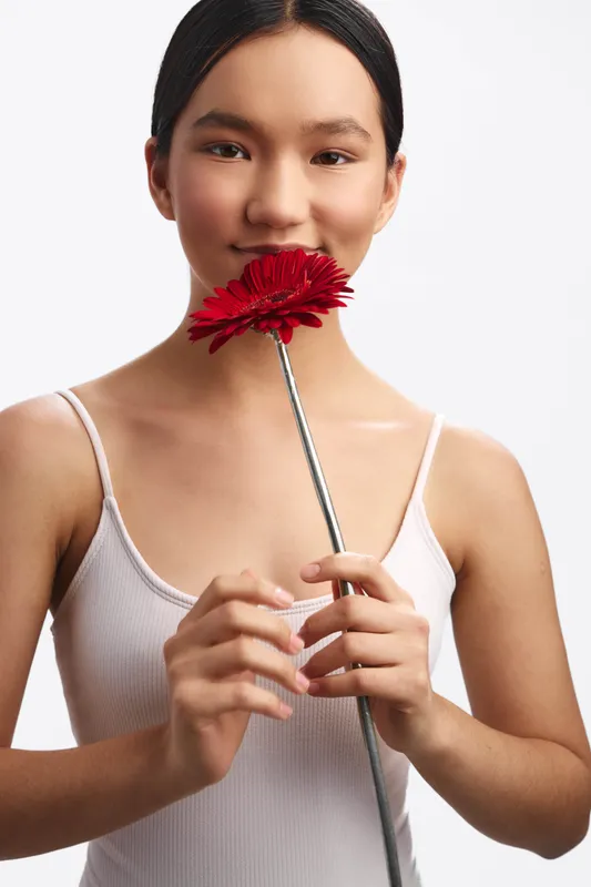 asian woman in a white tank top holds a red flower near her lips, creating a soft studio portrait with calm mood. bright, natural lighting highlights her serene expression and minimalist setting,