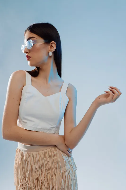 Young woman in white top and fringed skirt poses confidently against a clear sky. She wears large earrings and reflective sunglasses. Concept of woman beauty fashion and style.