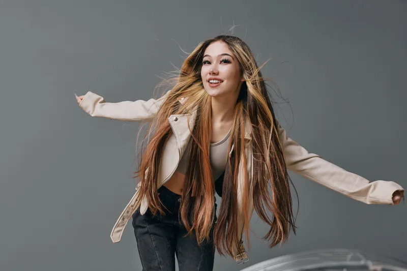 Young woman with long hair wearing casual outfit and jacket posing with arms wide open against plain gray background in studio for lifestyle and fashion concept.