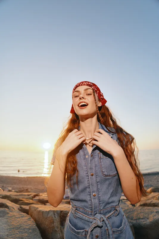 Lifestyle woman in modern boho-western denim street style wearing a red bandana, enjoying sunset by the beach with warm film color tones and natural light.
