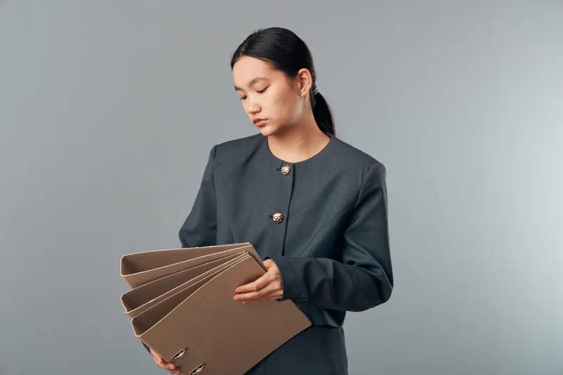 Young woman in dark blazer holding several brown folders indoors, looking down with focused expression against plain background. Businesswear, documents and office concept with calm atmosphere.