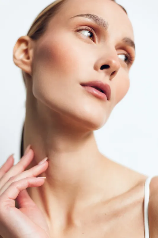 Close up of a young woman with flawless makeup looking to the side, soft lighting highlights her smooth skin and defined features, with a calm, introspective mood and subtle rosy tones.