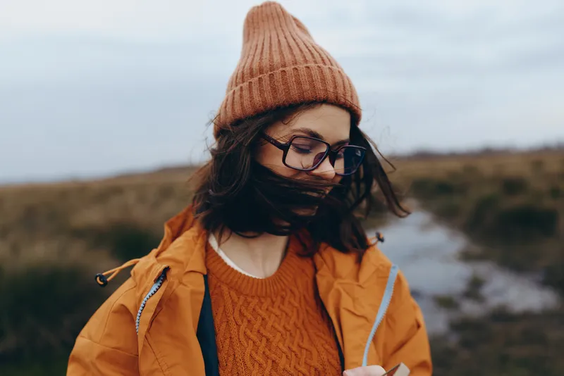 Woman lifestyle in winter outdoors, knit hat and glasses, orange coat over knit sweater, smiling while walking on a rural path, cloudy sky, fresh air, countryside hills