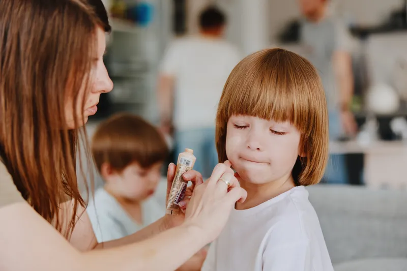 Caring woman applying makeup to her daughter while family interacts in the background, creating a warm and nurturing atmosphere at home.