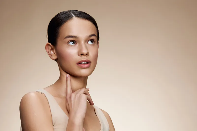 Thoughtful young woman with smooth skin, light makeup, and neutral attire, posing against a soft beige background, conveying beauty and elegance