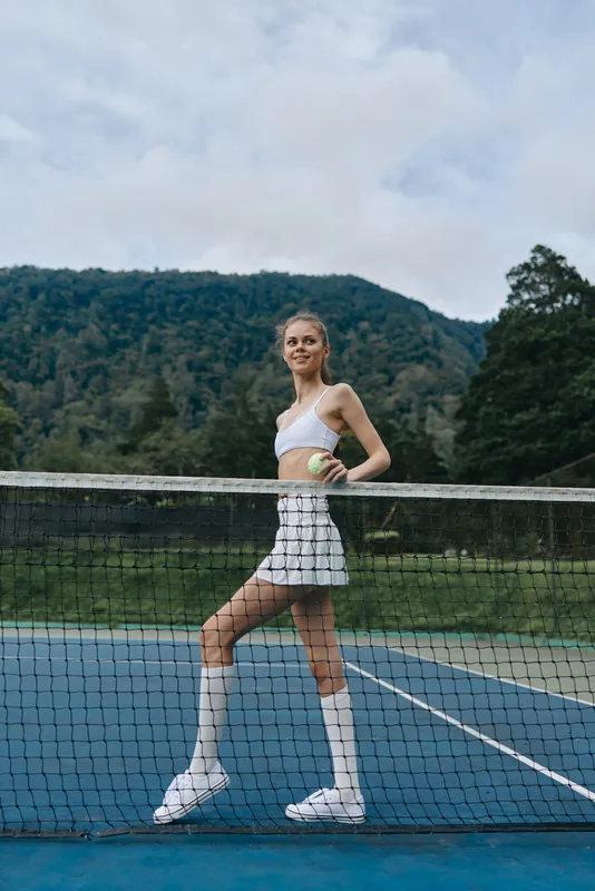 A young female athlete in a white tennis outfit poses confidently on a court with a tennis ball, showcasing her athleticism and determination against a natural background