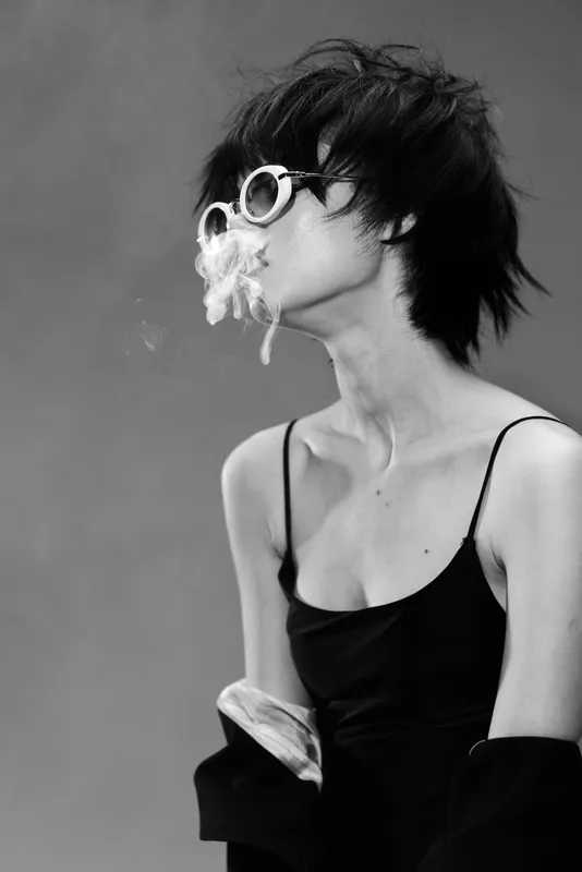 Black and white portrait of a young woman in sunglasses exhaling smoke with moody expression and messy hair style.