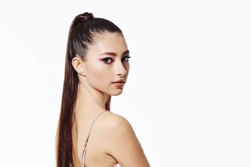 Young woman with sleek ponytail and bold eye makeup looks sideways against a white background fashion concept highlighting beauty and creative style expression.