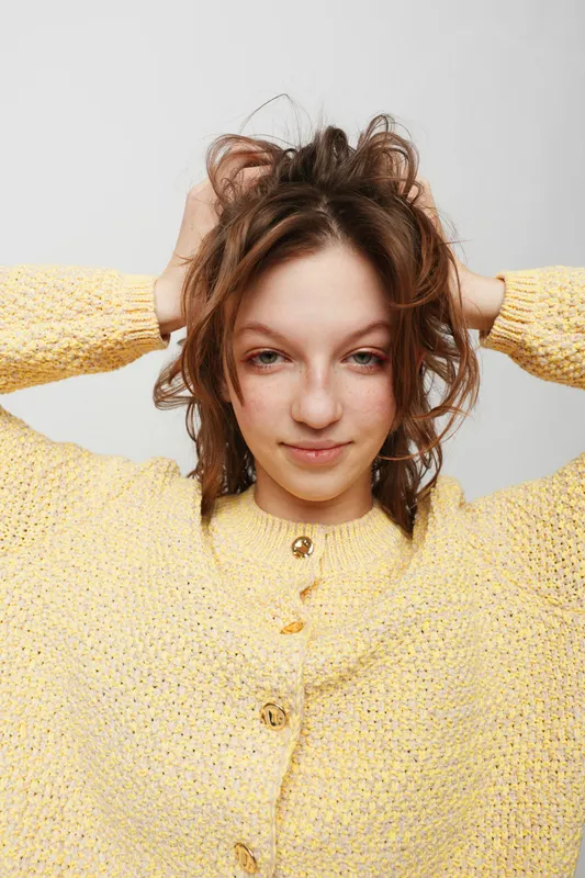 Young woman in yellow knitted sweater touches her messy hair, looking confident and relaxed. Studio portrait on white background with natural expression and casual style.