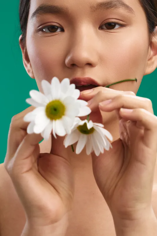 asian woman in a studio close up holds white daisies, soft gaze toward the camera, serene expression conveys freshness and beauty, minimal background highlights natural charm and delicate petals.