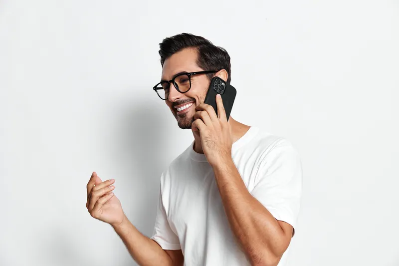 Full body smiling happy cheerful young man wearing glasses speaking on mobile phone while standing against plain light background. People lifestyle concept.