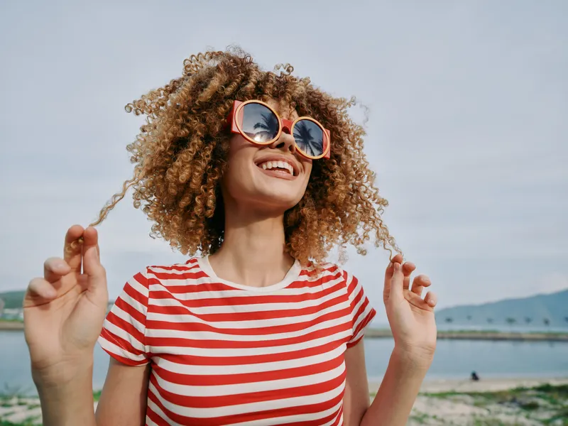 Bright portrait of a joyful woman with curly hair wearing a red striped tee and round sunglasses, outdoors by a lake, capturing a carefree summer mood and playful sunshine.