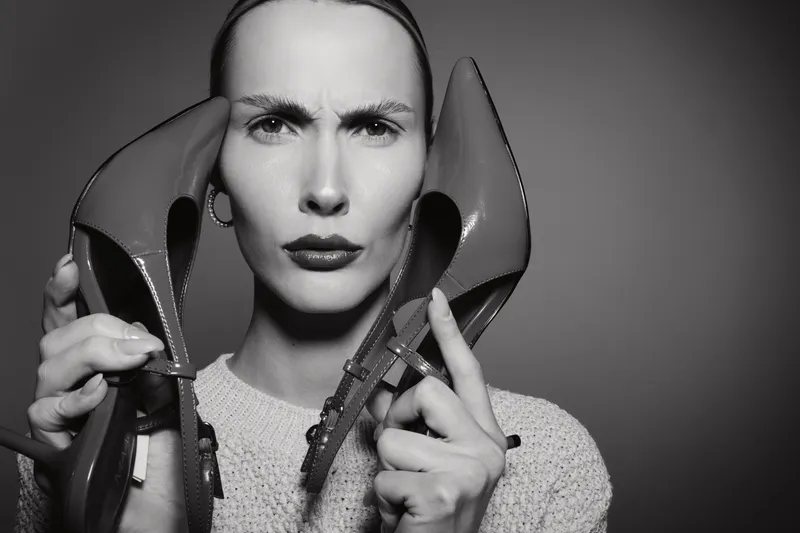 Serious white woman with pale skin holds high heel shoes near her face, showing frustration. Black and white studio portrait with sharp lighting. Concept for fashion and emotion.