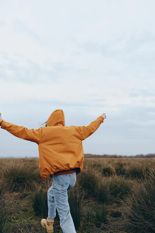 Woman outdoors in a countryside field, wearing yellow jacket with hood, walking forward with arms outstretched, joyful lifestyle moment, soft overcast light, natural grass background