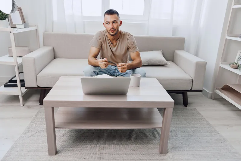 Focused man sitting on a couch, working on a laptop with a cup of coffee nearby, surrounded by a bright, modern interior that promotes productivity and creativity.