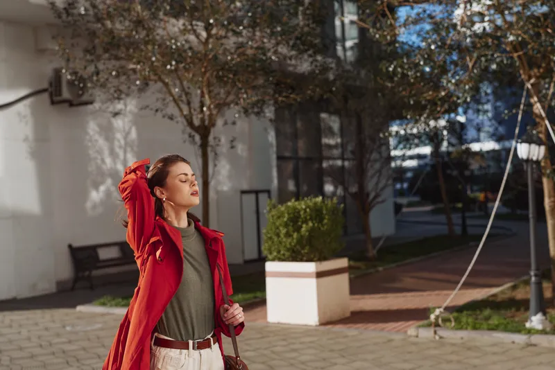 Young woman in a red coat enjoys a sunny autumn day outdoors with her eyes closed and hand resting on her head, feeling relaxed and peaceful in an urban park setting.