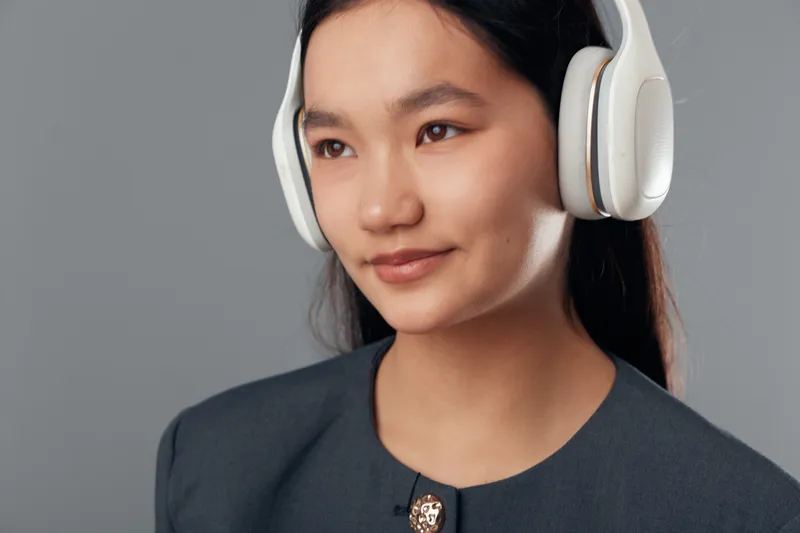 Young woman wearing wireless headphones and dark blazer smiling slightly looking away on gray background. Portrait technology person enjoying music or podcast with casual confident expression.