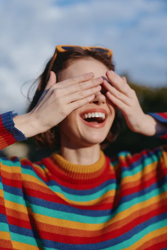 Woman smiling and laughing with hands covering eyes wearing colorful striped sweater and sunglasses on her head, playful outdoors portrait capturing joy and spontaneous expression.