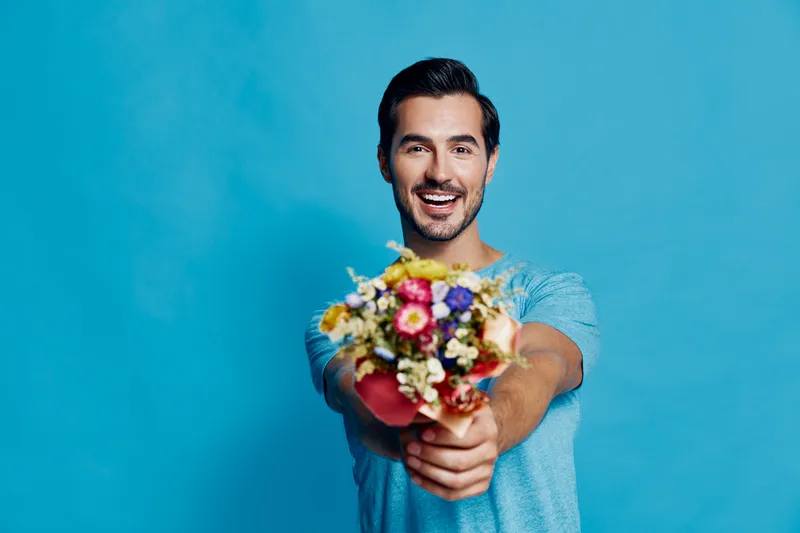 Happy cheerful young man smiling while holding a colorful bouquet of flowers in front of a bright blue background. Joyful expression and vibrant colors enhance the mood.