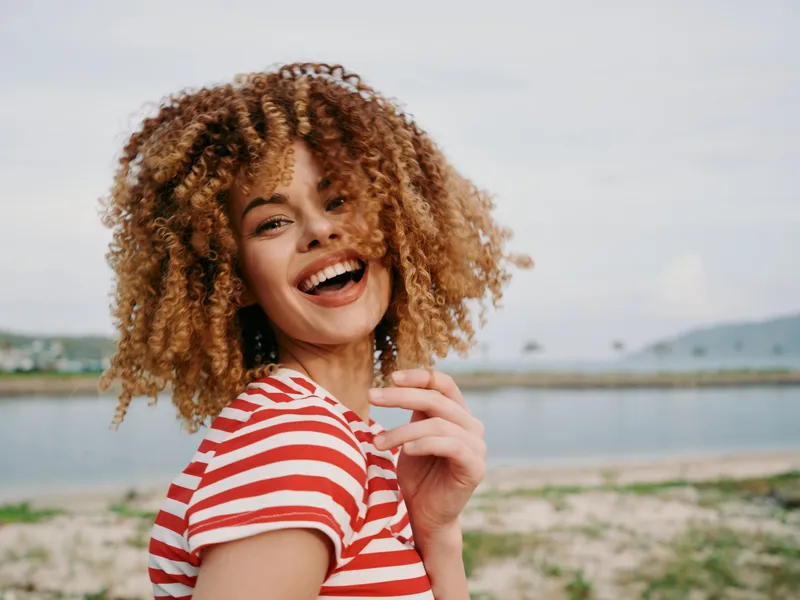 Joyful woman with curly hair smiles toward the camera on a sunny day, wearing a striped red and white shirt, with a lake and beach backdrop and a lively, candid vibe.