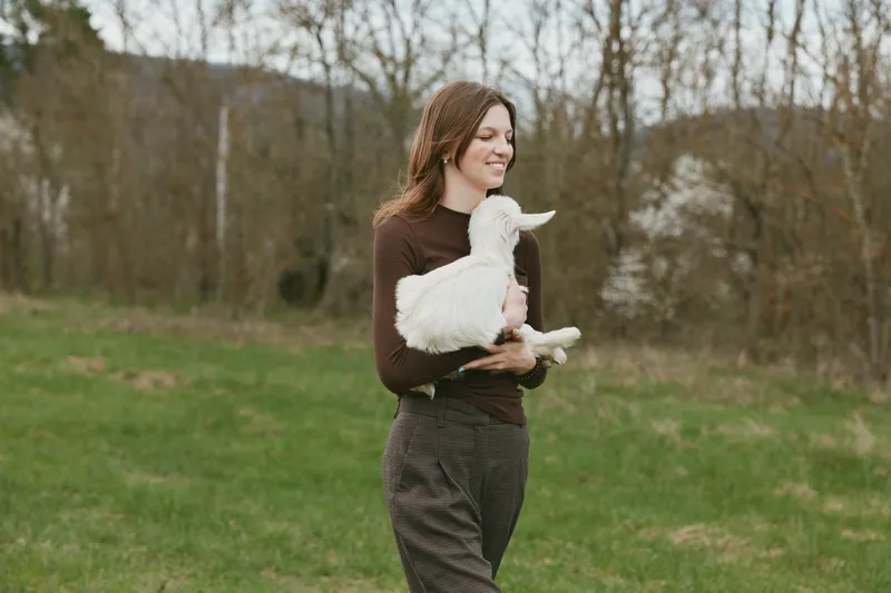 Smiling woman holds a goat kid in a grassy field, enjoying an outdoors moment with a calm rural feel, highlighting animal human bond and nature.