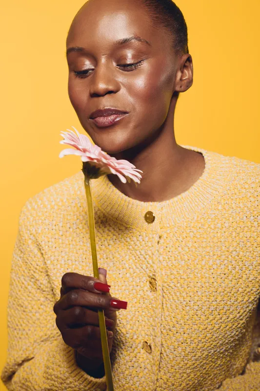 Young African woman with dark skin wearing yellow knitted sweater holds pink flower close to face, eyes closed, smiling gently. Studio portrait on yellow background, concept of beauty and calm.