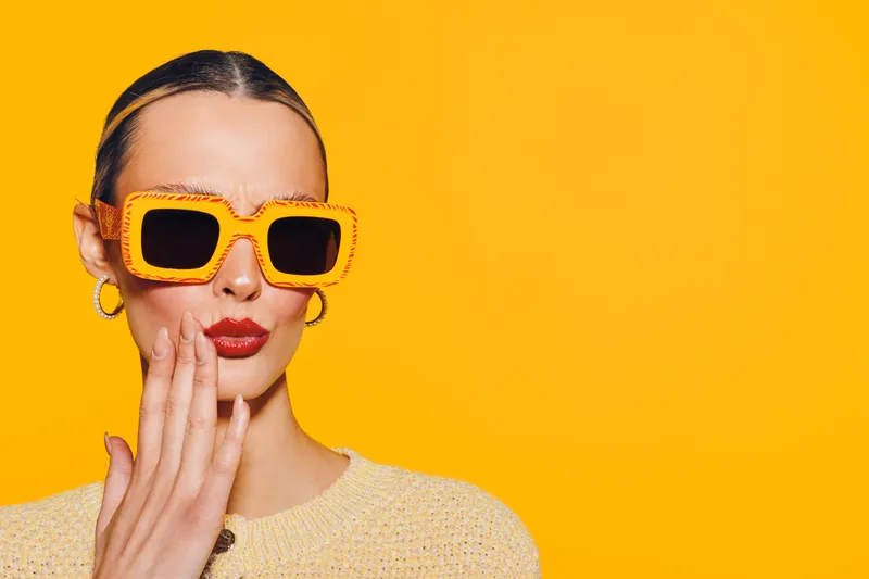 Stylish woman with slicked hair wearing large orange sunglasses and red lipstick poses against bright yellow background. Fashion concept with bold colors and confident expression.