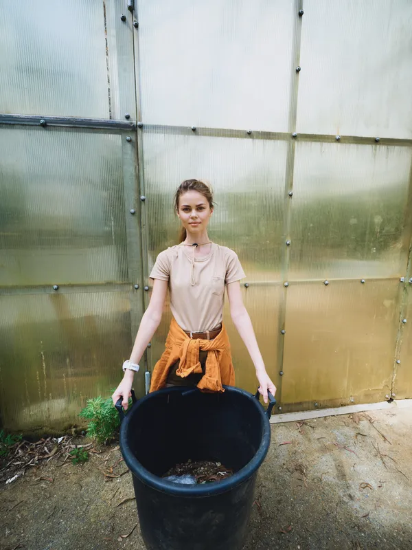 Woman in greenhouse gardening with a large black bin of soil and compost, plants and seedlings nearby; young female gardener with sweater tied at waist doing potting and watering.