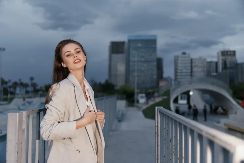 confident young woman in beige blazer standing outdoors in urban cityscape with moody sky, modern architecture, and metal railings