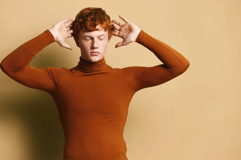Young man with red hair in brown turtleneck poses with closed eyes and hands near head in minimal studio setting with warm beige background and soft lighting.